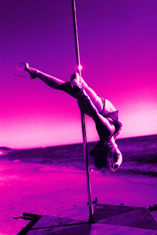 A pole dancer holding an inverted split on the pole by the water, in a pink and violet duotone