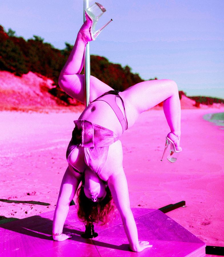 Rose in a handstand with her legs hooked around the pole, on a mat on the beach, in pink tones