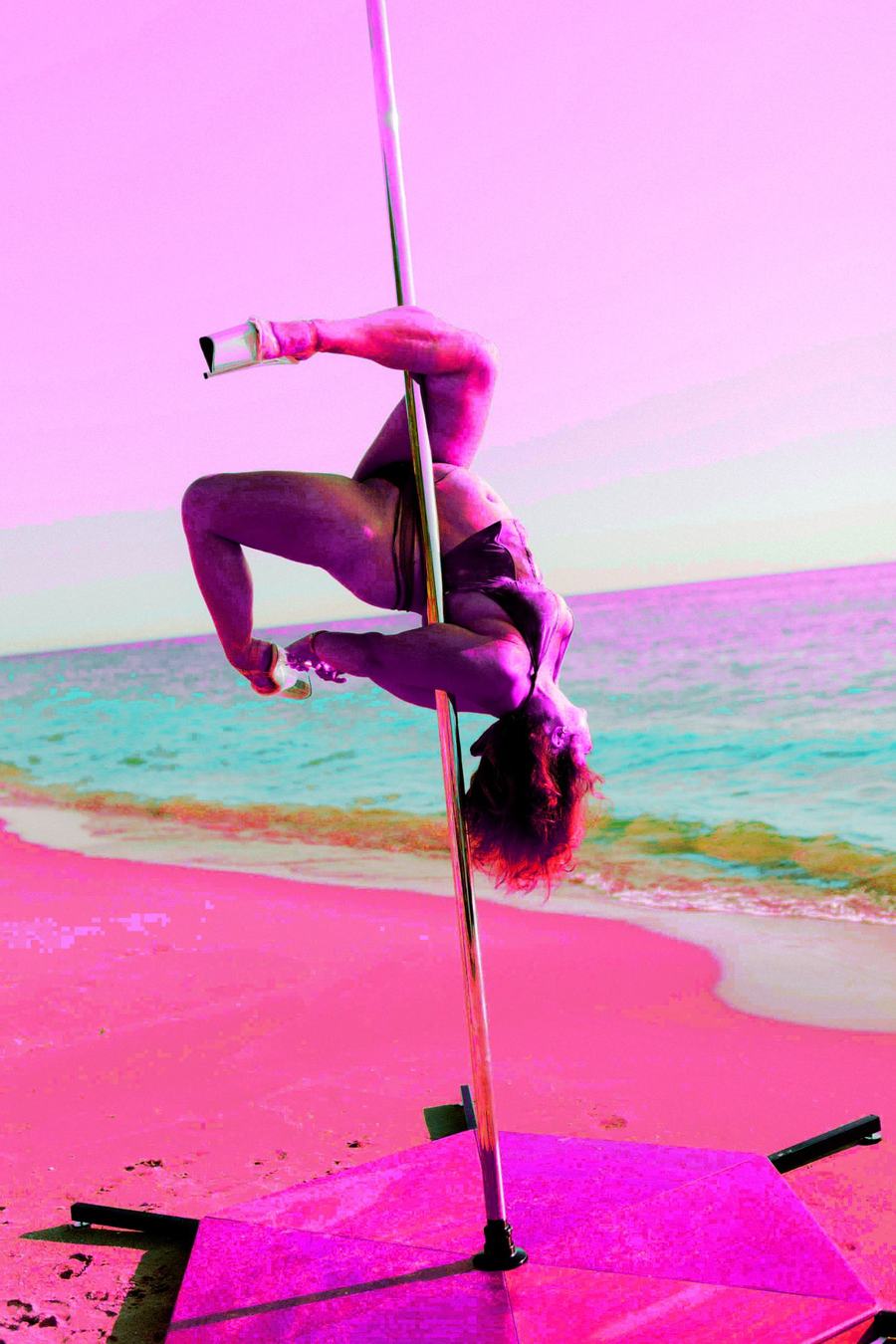 A pole dancer holding an inverted pose on a pole set up on the beach, tinted neon pink