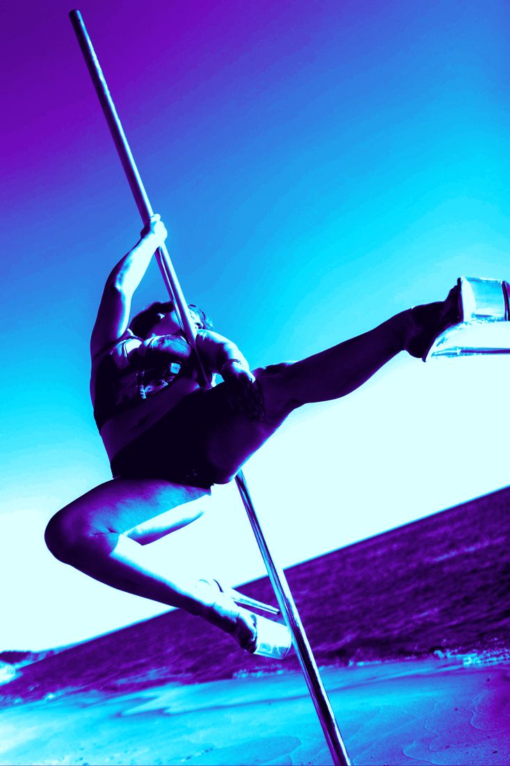 A pole dancer mid-kick on an outdoor pole on the beach, in a cyan and violet duotone